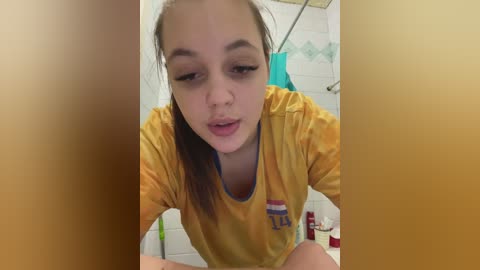 Media: A video shows a young woman with light skin and straight brown hair, wearing a yellow shirt, leaning forward in a tiled bathroom with a white sink and green soap dispenser.