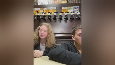 Media: Video of two Caucasian teenagers with curly hair, one smiling, sitting at a table in a restaurant. Background includes a wall with plastic figurines and a menu board.