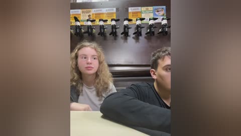 Media: Video of two teenagers in a dimly lit, wood-paneled room. A blonde girl with curly hair, wearing a white shirt, sits beside a boy with short brown hair in a black shirt. Black plastic figurines hang from the wall above.