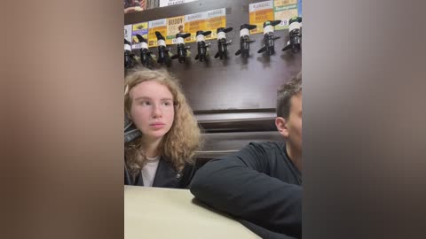 Media: Video of a young woman with long, curly blonde hair, wearing a black jacket, seated at a table in a dimly lit fast-food restaurant, surrounded by rows of black and yellow soda machines.