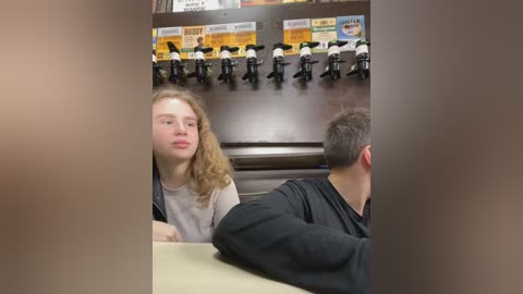 Media: Video of a young woman with curly hair and a light complexion, wearing a gray shirt, sitting at a table in a dimly lit restaurant. Behind her, a row of black mannequins dressed in white shirts and black pants, with a yellow sign above.