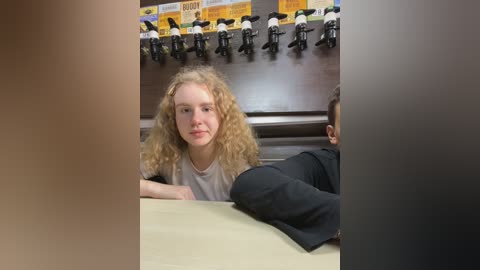 Media: Video of a young girl with curly blonde hair, wearing a white shirt, sitting at a table in a studio with black microphones and a yellow banner in the background.