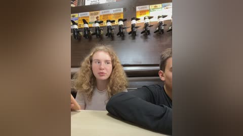 Media: Video of a young woman with curly blonde hair and a white t-shirt, seated at a table in a fast-food restaurant, surrounded by black and white Lego figures mounted on a wooden wall.