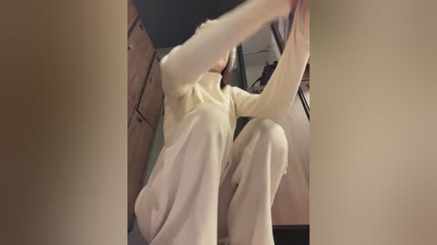 Media: A video of a person in a beige sweater and pants, crouching in a dimly lit room with wooden cabinets, a mirror, and a blurred background.