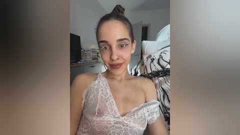 Media: Video of a young woman with light skin, brown hair in a bun, wearing a lacy white top, sitting on a bed with a zebra-print pillow, in a dimly lit bedroom.