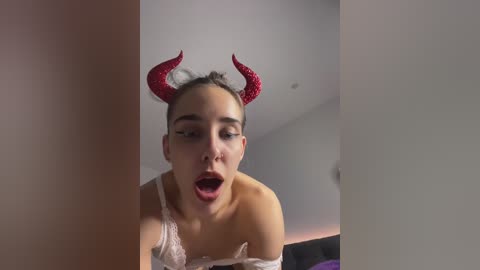 Media: Video of a woman with light skin, wearing red devil horns and a white lace bra, mouth open in an expression of ecstasy, in a dimly lit room.