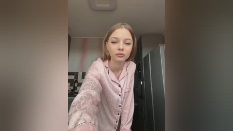 A video of a young Caucasian woman with straight blonde hair, wearing a light pink satin robe, taken from a selfie perspective. The background features a modern kitchen with grey and black tiles.