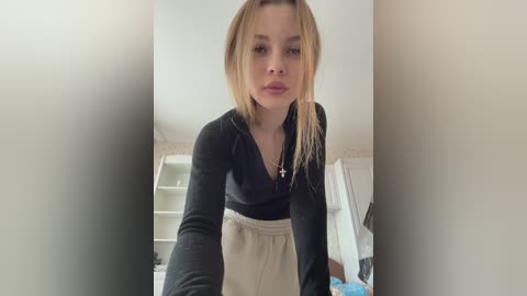 Media: A video of a young woman with long, straight blonde hair, fair skin, and wearing a black V-neck sweater and beige sweatpants, standing in a bright, minimalistic room with white walls and a bookshelf.