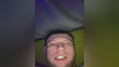 A close-up video of a young, white man with short brown hair and a beard, smiling, lying on a green pillow. The background is dimly lit with purple and green hues, suggesting a nighttime setting.