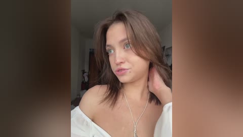 Video of a young woman with shoulder-length brown hair, fair skin, and light makeup, wearing a white off-shoulder top and a silver necklace, standing in a blurred bedroom with a closet and hanging clothes in the background.