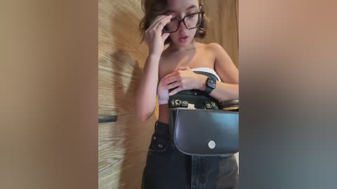 Media: Video of a young woman with glasses, medium build, wearing a white strapless top, black high-waisted pants, and a black VR headset. She is adjusting her glasses and holding a black handbag. Background shows wooden walls and a blurry doorway.