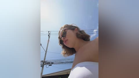 Media: A video of a Caucasian woman with short, wavy blonde hair, wearing white sunglasses and a white off-shoulder top, leaning against a light blue sky with power lines visible in the background.