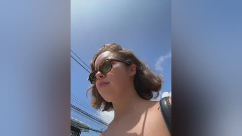 Media: Video of a fair-skinned woman with shoulder-length brown hair, wearing aviator sunglasses, standing outdoors against a clear blue sky. She appears to be smiling slightly.