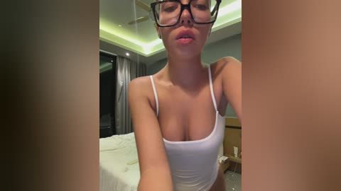 Media: A video of a young Asian woman with light skin, wearing glasses, a white tank top that reveals her ample cleavage, and black high-waisted pants, posing in a modern bedroom with soft green lighting and a neatly made bed.