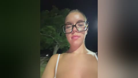 Media: Video of a Caucasian woman with light skin and blonde hair, wearing black-framed glasses and a white top, standing outdoors at night with a blurred tree in the background.