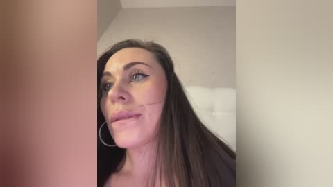 A video of a fair-skinned, brunette woman with long hair, wearing hoop earrings, in a neutral-toned room with a white headboard visible in the background.