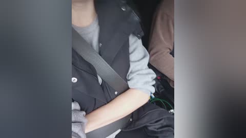 A video of a person in a car seat, wearing a black jacket over a gray shirt, with a shoulder strap and a green item on the right side.