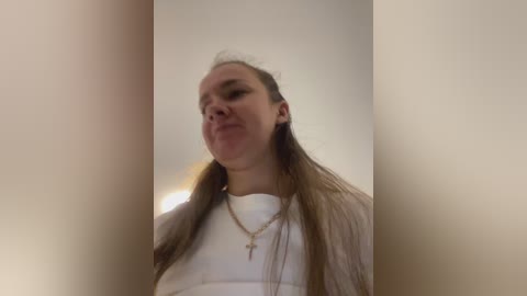 Video of a young woman with long, straight brown hair, wearing a white top, and a cross necklace, making a pouty face, taken from a low angle.