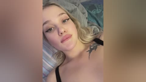 Media: Video of a young Caucasian woman with blonde wavy hair, wearing black lingerie, lying on a bed with patterned pillows. She has a tattoo on her right shoulder.