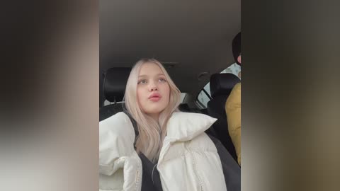 Media: A video captures a young woman with long, straight blonde hair, wearing a white jacket and black top, seated in a car with dark leather seats.