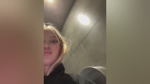 Media: A video of a young woman with blonde hair, light skin, and a neutral expression, taken in a bathroom with beige tiled walls and a ceiling light.