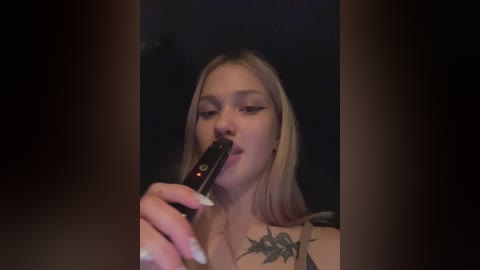 Media: Video of a young blonde woman with light skin, wearing a black top, smoking a black cigarette, with a large black tattoo on her right shoulder.