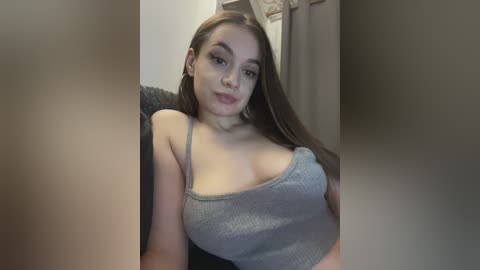 Video of a young Caucasian woman with light skin and long brown hair, wearing a gray ribbed tank top, showing cleavage. She sits on a dark couch with a beige curtain in the background.