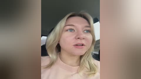 Video of a Caucasian woman with fair skin and shoulder-length blonde hair, wearing a light pink shirt, sitting in a car. The background is blurred, and the image has a soft focus.