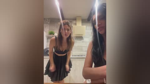 Video of two young women in a modern kitchen; one in black lingerie, the other in a black dress, both leaning forward with playful expressions.