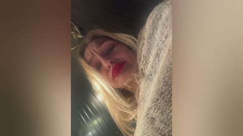 Media: Video of a blonde woman with fair skin, closed eyes, and red lipstick, wearing a textured, light gray sweater, seen from a low angle.