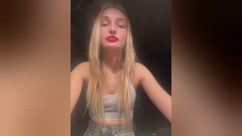 Media: Video of a blonde woman with fair skin and red lipstick, wearing a white tank top and high-waisted jeans, looking serene against a dark, blurred background.