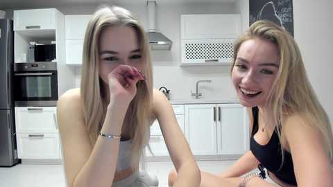 Media: Video of two smiling young women with fair skin and long blonde hair, one in a white top, the other in a black sports bra, sitting in a modern kitchen with white cabinets, stainless steel appliances, and a chalkboard wall.