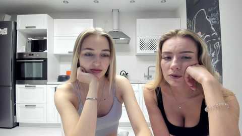 Media: Video of two young Caucasian women with blonde hair, one in a white tank top and the other in a black tank top, sitting on a kitchen counter, looking contemplative, in a modern, white kitchen with stainless steel appliances.