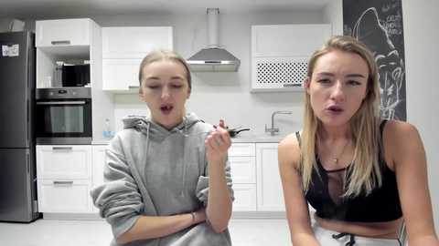 Media: Video of two blonde, fair-skinned women in a modern kitchen; one wears a gray hoodie, the other a black bralette. Both hold forks. Background includes white cabinets, stainless steel appliances, and a chalkboard wall.