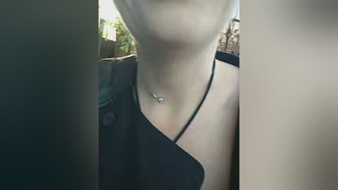 A video captures the lower face and upper chest of a light-skinned woman wearing a black, sleeveless top. She has a silver necklace with a small pendant. The background is blurry, suggesting an outdoor setting with trees.