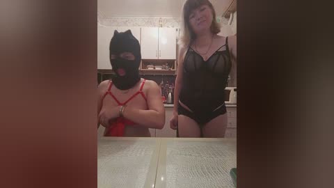 Video of two women, one in a black leather mask and red lingerie, the other in a sheer black bodysuit, in a kitchen.