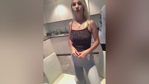 Media: Video of a slender, fair-skinned blonde woman with straight hair, wearing a dark spaghetti-strap top and light blue leggings, standing in a modern kitchen with white cabinets and gray countertops.