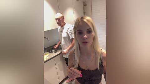 Media: Video of a blonde woman with pink nails and a black top, standing in a modern kitchen with white cabinets and a sink. An elderly man with glasses and a white shirt is washing dishes behind her.