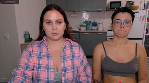 Media: Video of two women in a kitchen: one in a pink plaid shirt, the other in a grey sports bra, both with neutral expressions.