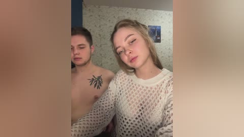 Video of two young, shirtless men and a woman in a dimly lit room. The woman, with blonde hair, wears a white, fishnet sweater, while the men have tattoos.