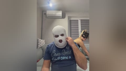 Video of a man in a white balaclava mask, blue \"Lady Rod\" T-shirt, and black mask, holding a black mask; a blonde woman in a black top, also masked, is in the background.