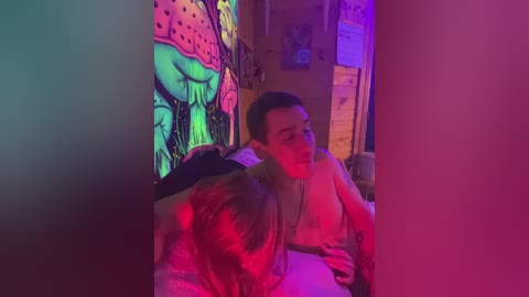 Video of a dimly lit bedroom with vibrant neon lighting, showing a young man and woman sitting on a bed, both shirtless, under a colorful, psychedelic mural.