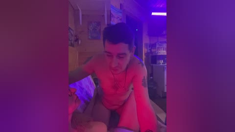 Video of a young, nude man with a slender build and short dark hair, kneeling in a dimly lit bedroom with purple and red lighting.