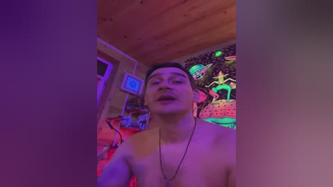 A video of a shirtless young man with short dark hair, wearing a necklace, standing in a colorful, dimly lit room with a mural of space-themed art on the wall.