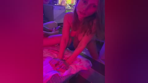 Video of a nude woman with fair skin and long hair, leaning over a bed with pink sheets, in a dimly lit room with purple and pink lighting.