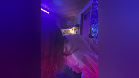 Video of a young woman with long, brown hair bent over, wearing a white shirt, in a dimly lit, purple-lit room with a fish tank and wooden ceiling.