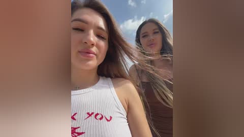 Media: Video of two young women with long brown hair, one in a white ribbed crop top with red text, the other in a beige top, outdoors with a clear blue sky.