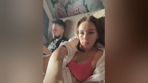 A video shows a bearded man in a black shirt, sitting on a couch, and a woman with light brown hair, wearing a pink bra, sitting beside him, both looking introspective.