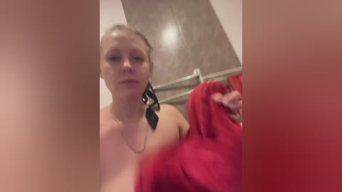 A video of a topless woman with light skin and auburn hair, wearing a black necklace with a feather pendant, holding a red towel in a bathroom with beige tiles.
