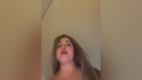 A blurry video of a plus-sized woman with long, wavy brown hair, wearing dark lipstick, captured from a low angle, against a beige wall.